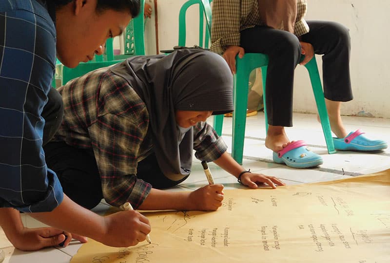 A woman in a hijab and another team member write on a large piece of paper on the floor