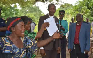 A woman smiles holding up a piece of paper, with officials in the background