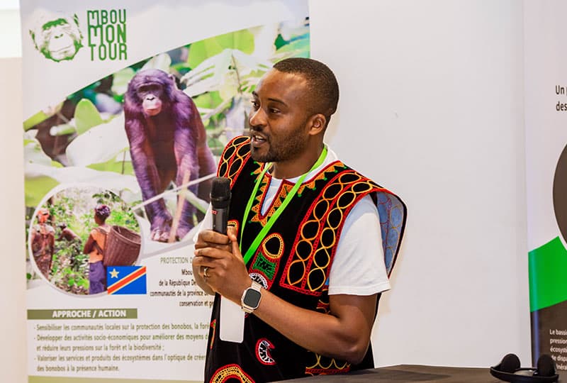 A person speaks into a microphone with a bonobo poster in the background