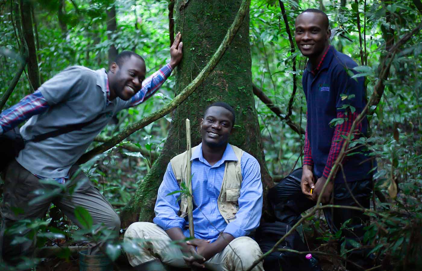 Herp Ghana Conservation lead, Caleb Ofori Boateng crouched down, smiling and leaning against a tree, with two colleagues in forest